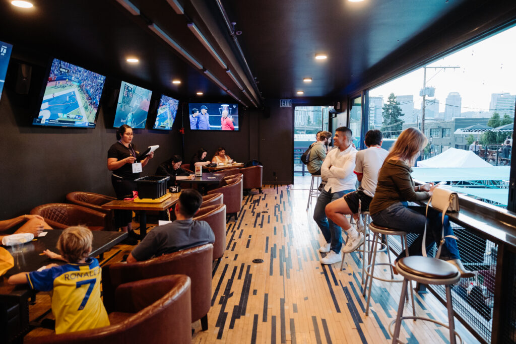 Indoor bar lounge at WonderLove with guests watching sports on multiple wall-mounted TVs, seated at leather chairs and window bar seating.