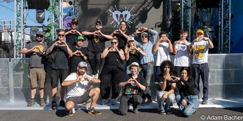 WonderLove team members posing together on an outdoor stage in daylight, smiling and forming heart shapes with their hands in front of a large LED screen and stage lighting rig.