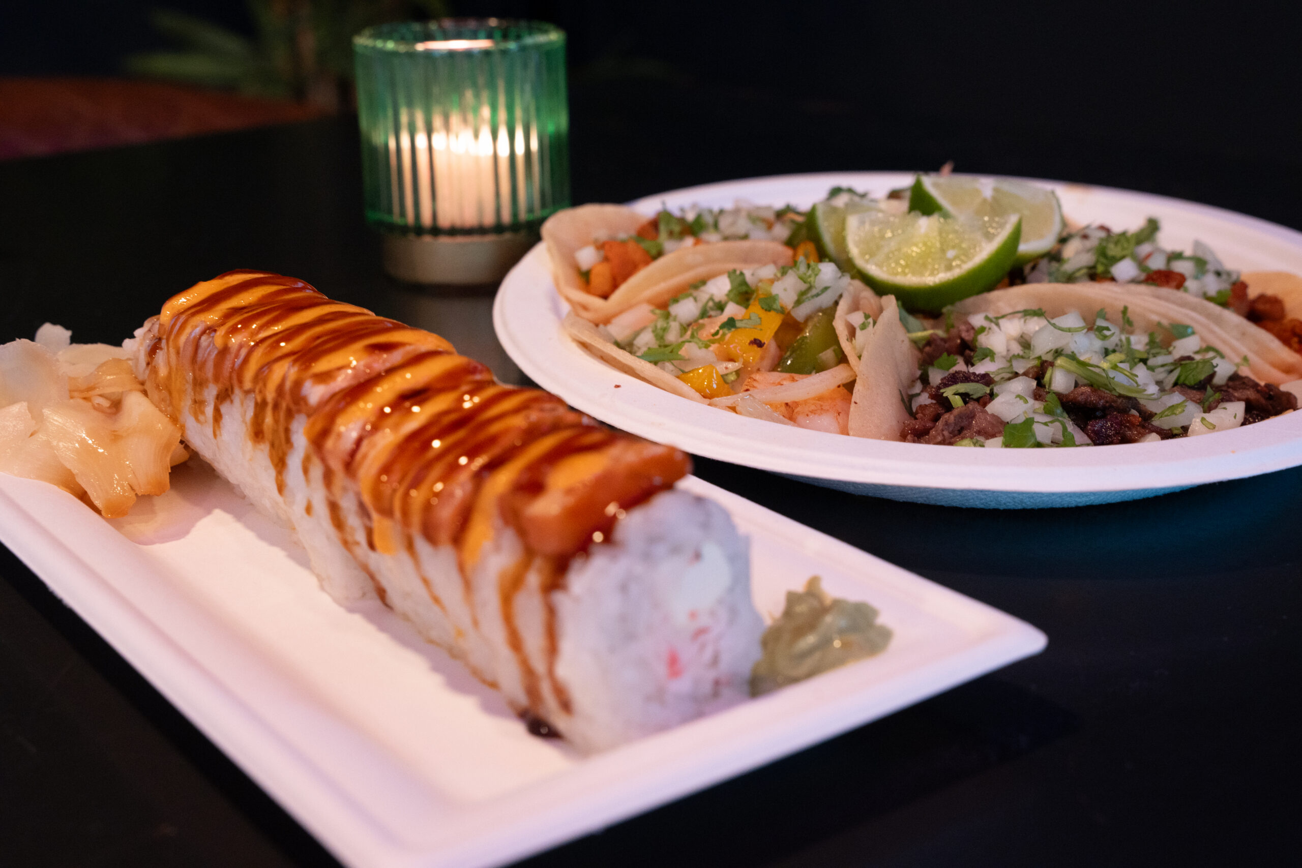 A sushi roll drizzled with sauce on a white plate sits in the foreground, with street-style tacos topped with cilantro, onion, and lime on a plate behind it.