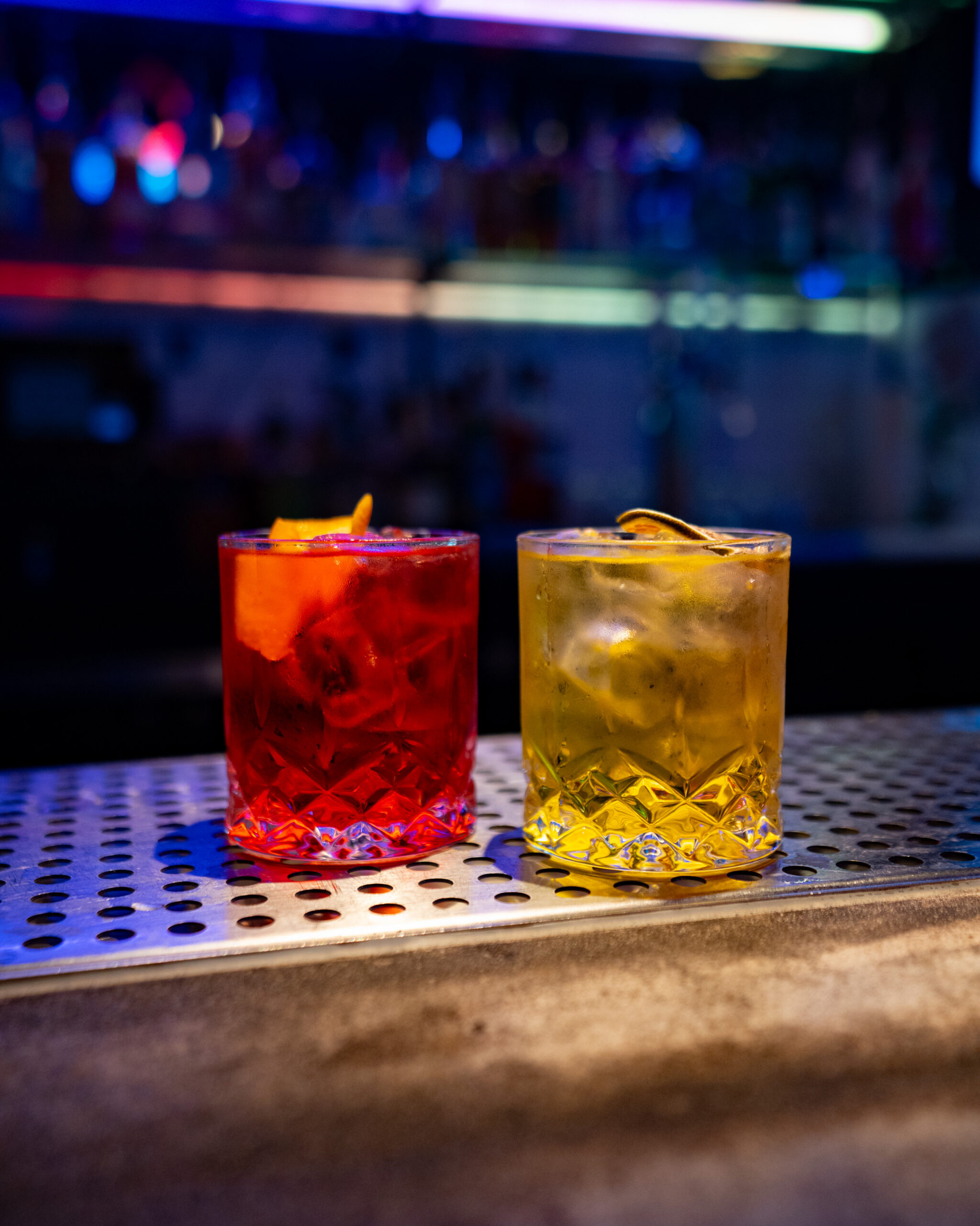 A low-angle photograph taken on a metal bar counter shows two custom-made cocktails. On the left is a vibrant red drink with an orange twist garnish. On the right is a golden-yellow drink garnished with a dehydrated citrus wheel. Both are served in patterned rocks glasses with ice, illuminated by colorful, blurry neon bar lights in the background.
